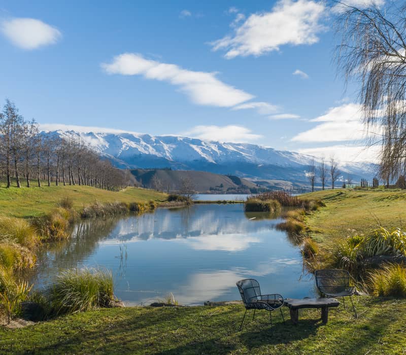 Two black mesh outdoor chairs and a small wooden table sit on a grassy bank overlooking a calm pond that reflects snow-capped mountains and a blue sky at Cloudy Bay vineyard.