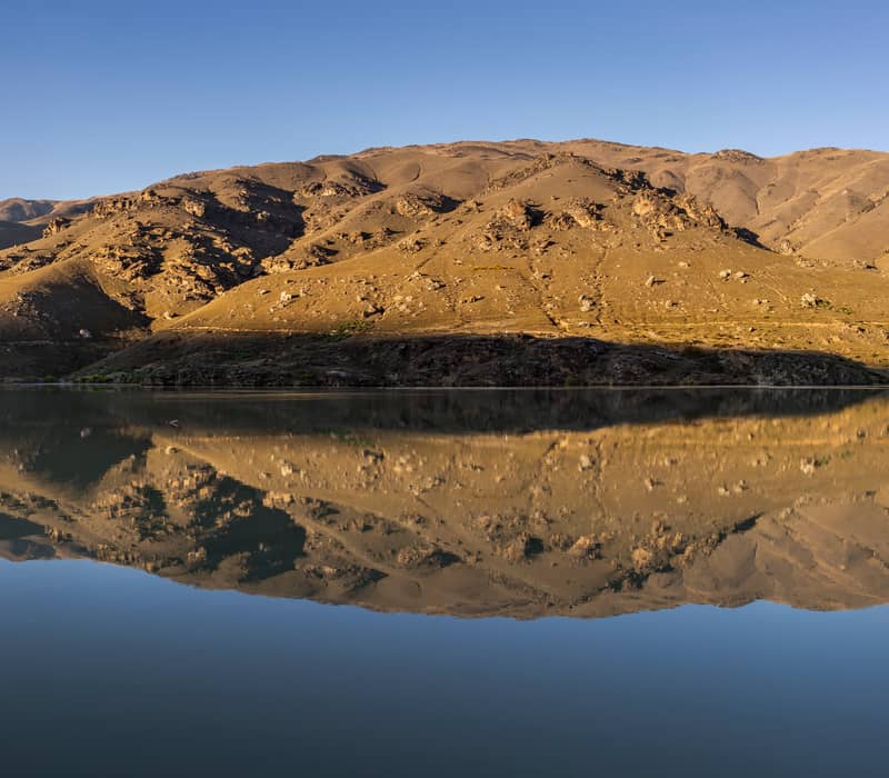 A wide panoramic view of Lake Dunstan in Central Otago, showing a perfect mirror reflection of sun-drenched, rocky brown mountains under a clear blue sky.