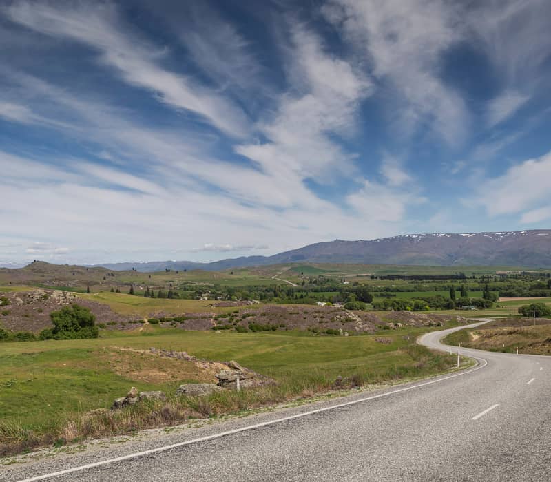 A winding two-lane highway curves through a vast landscape of green fields, rocky schist outcrops, and distant purple mountains under a dramatic blue sky with wispy white clouds.