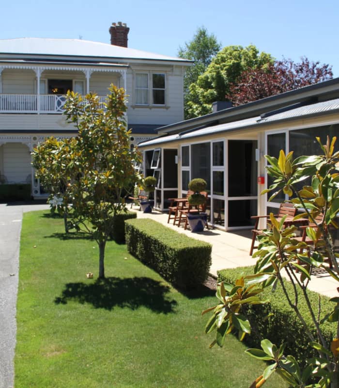 Two-story manor building with garden, flowering plants, and benches outside on a sunny day. Credit: Merivale Manor - Christchurch