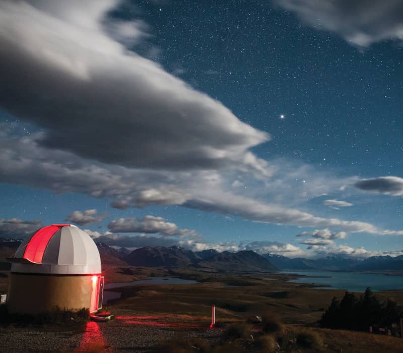 Observatory dome with red light under a starry sky and mountains by a lake at night. Credit: Dark Sky Project