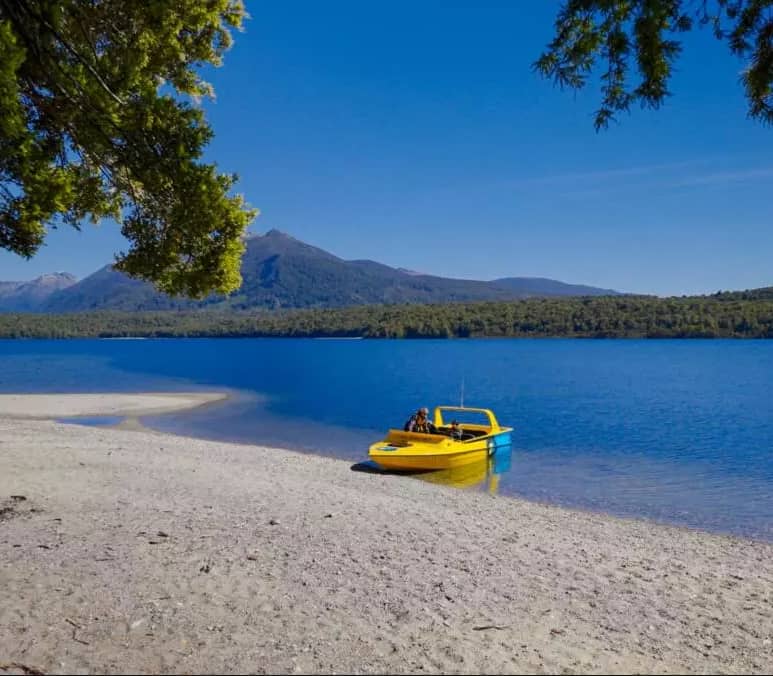Yellow boat on a lake with sandy beach, forest, and mountains under blue sky. Credit: Fiordland Jet