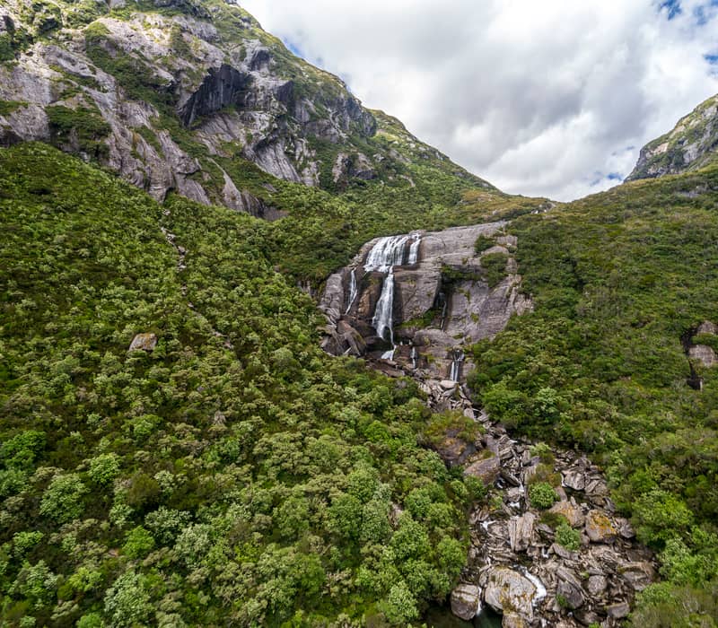 A wide waterfall cascades down a steep, rocky mountain face surrounded by dense green native bush in the Haast Pass.