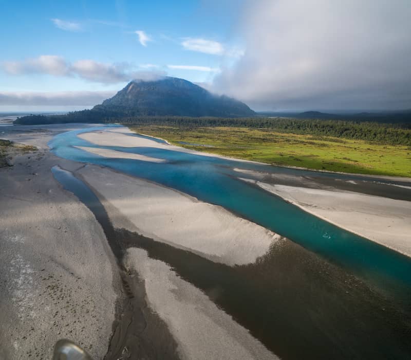 An aerial view of the Haast River’s turquoise braided channels flowing through wide shingle banks toward a forested mountain under a soft, morning sky.