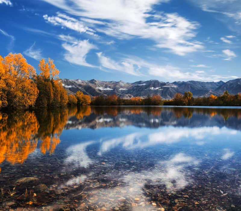 A wide landscape of Lake Ruataniwha in autumn, featuring brilliant orange trees and snow-dusted mountains reflected in the still, clear water under a blue sky with streaky white clouds.