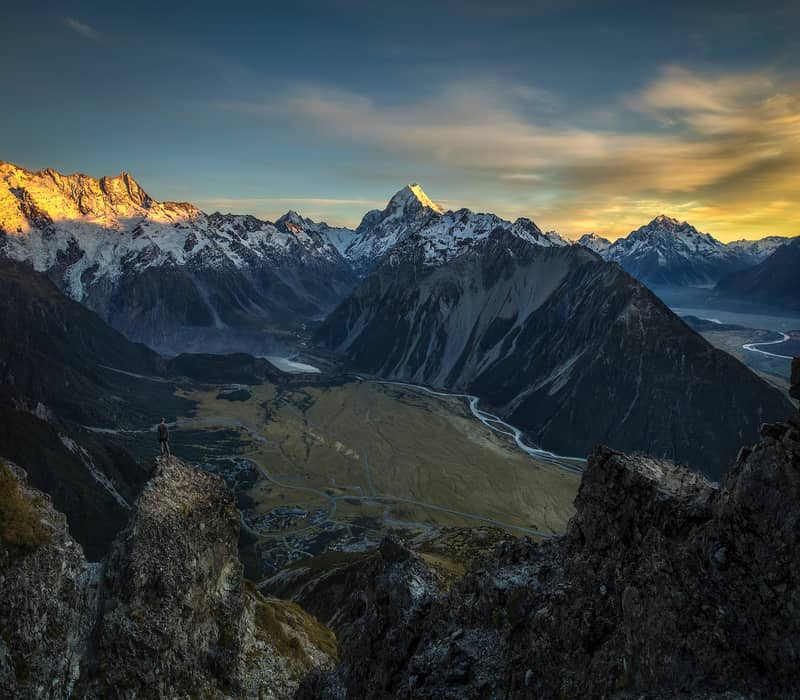 A high-altitude view of a person standing on a rocky outcrop overlooking a vast glacial valley, with the sun-drenched peak of Aoraki / Mount Cook in the distance under a golden sky.