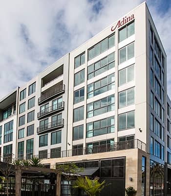 Modern multi-story hotel building with glass windows and balconies under a partly cloudy sky. Credit: Adina Britomart Hotel Auckland