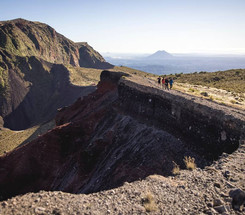 A group of four hikers walks along the narrow, rocky rim of a massive volcanic crater on Mount Tarawera, with deep red and purple chasm walls falling away to the left.