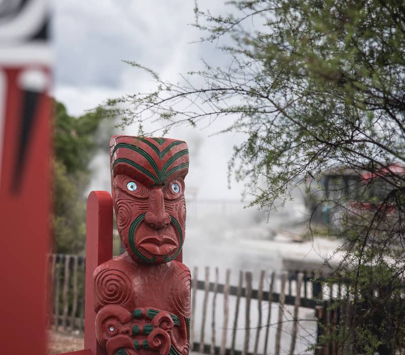 A close-up of a traditional red Māori wood carving with iridescent pawa shell eyes, standing in front of a steaming geothermal field and native manuka trees.