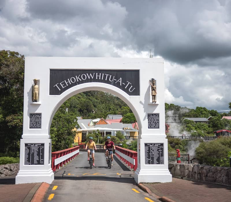 Two cyclists ride through the historic white "Te Hokowhitu-a-Tu" memorial archway into Whakarewarewa Living Māori Village, with geothermal steam visible in the background.
