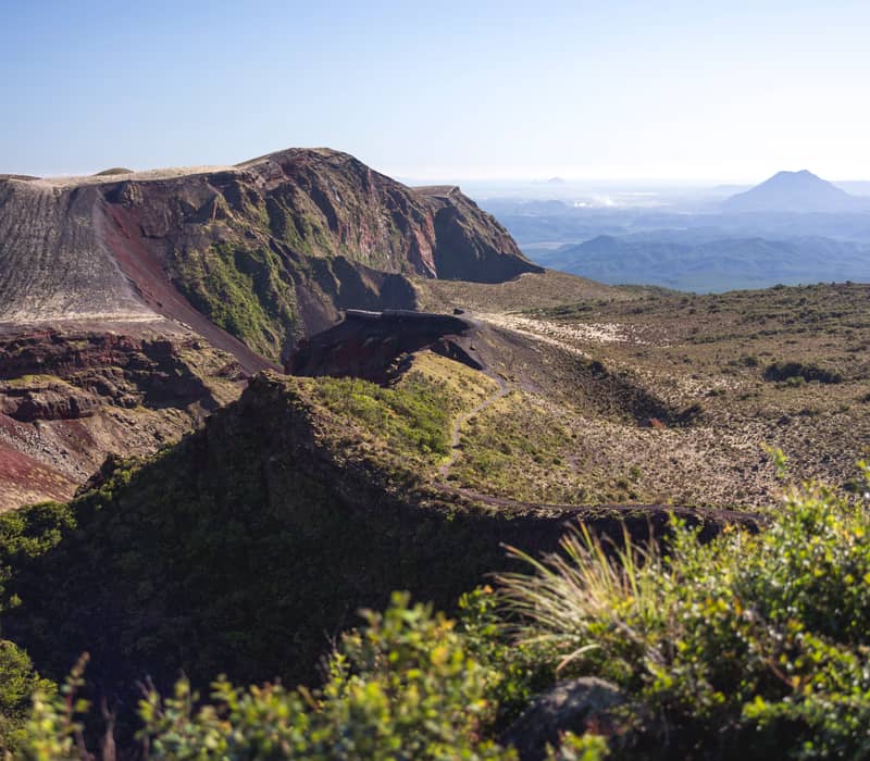 An expansive view of the deep, reddish-brown volcanic crater of Mount Tarawera in Rotorua, with a hiking trail winding along the rim and distant mountains under a clear sky.