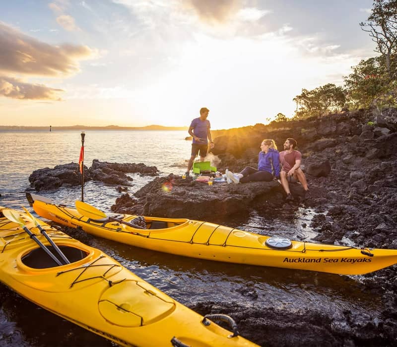 Two yellow kayaks on water near rocky shore with couple enjoying a picnic at sunset. Credit: Auckland Sea Kayaks