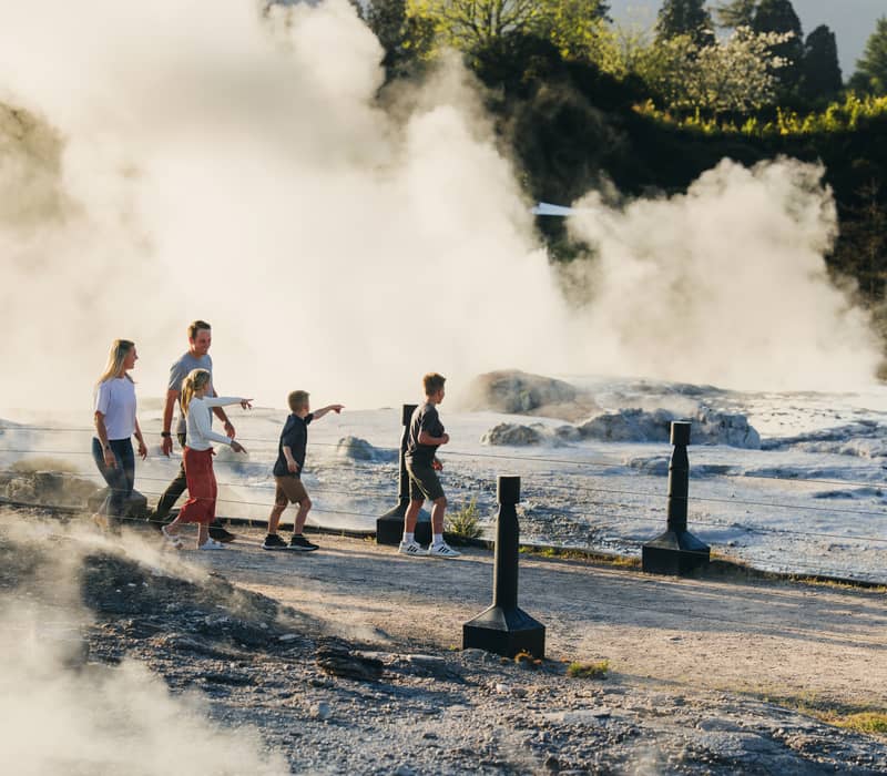 Family walking along a pathway with geothermal steam rising nearby in a green outdoor area. Credit: Te Puia