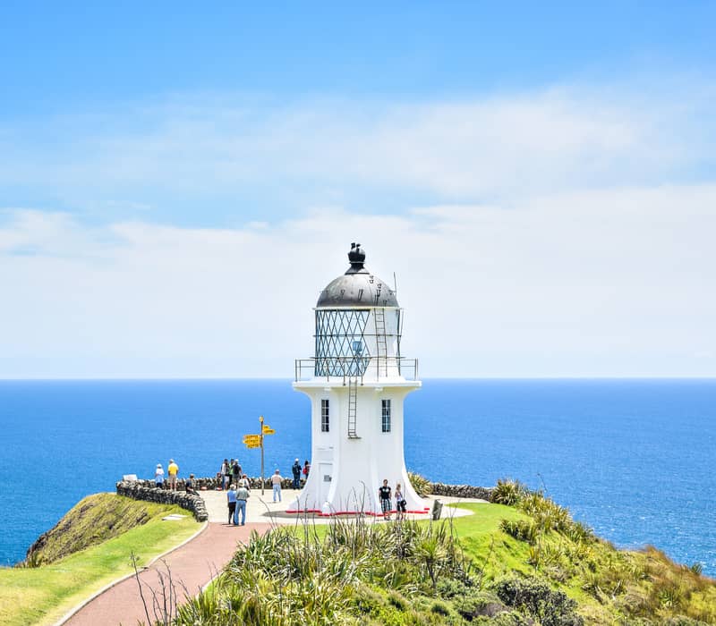 White lighthouse on cliff edge with people and vast blue ocean under a partly cloudy sky