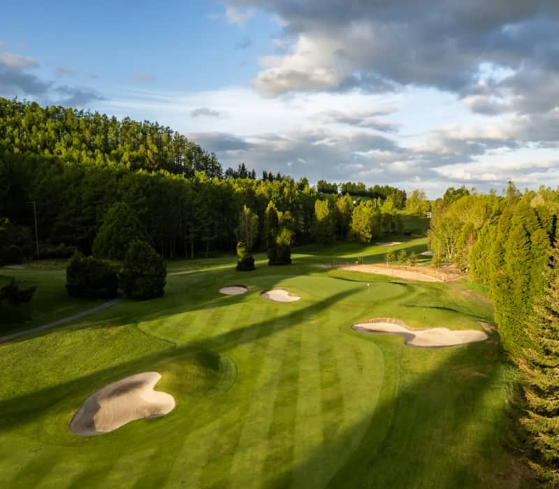 An aerial view of the lush green fairway and sand bunkers of Wairakei Golf + Sanctuary under a partly cloudy sky at sunset.