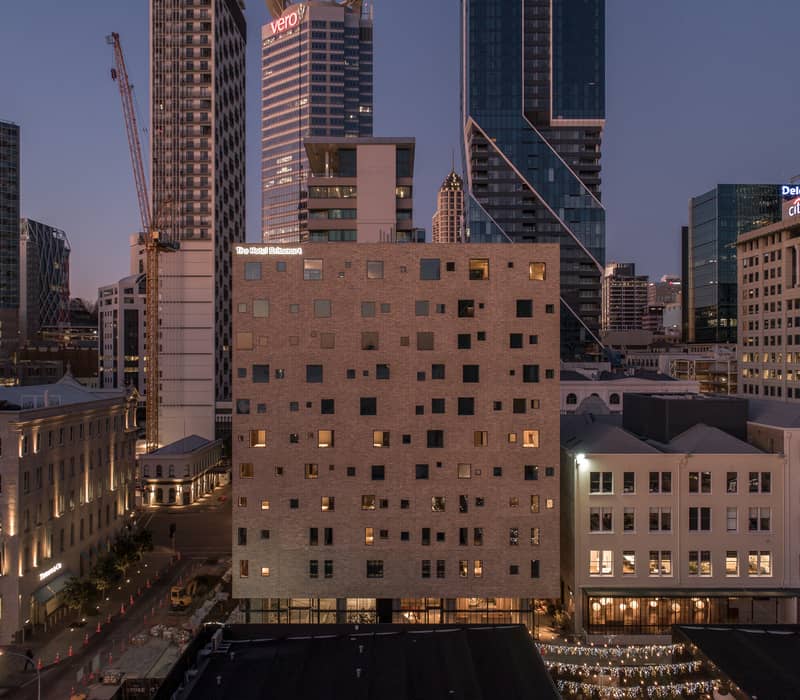 The Hotel Britomart building lit up at dusk in a city with tall buildings and cranes. Credit: Hotel Britomart