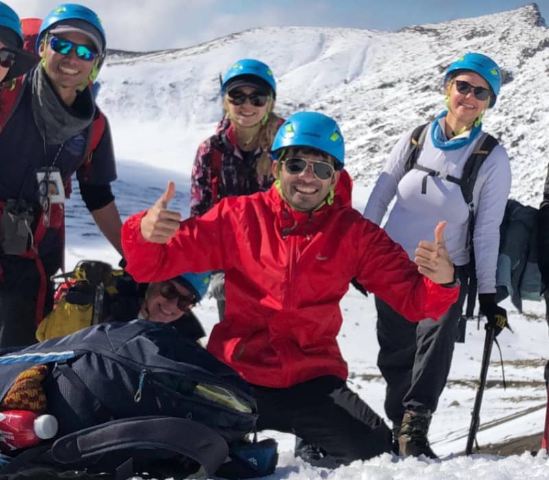 A smiling group of seven hikers in blue helmets and sunglasses posing on a snowy mountainside with rocky peaks in the background. Credit: Adrift Tongariro