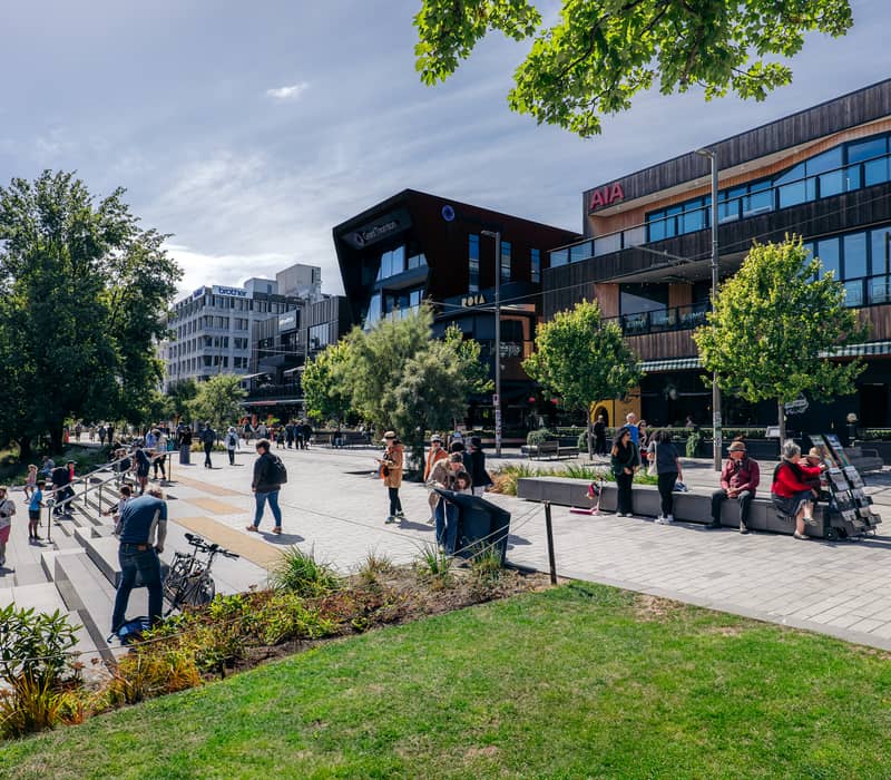 A busy midday scene on Oxford Terrace in Christchurch, showing people walking past modern office buildings and the riverside promenade.