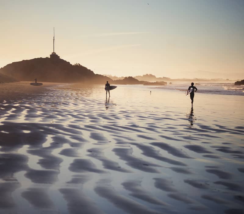 Silhouetted surfers walking on rippled wet sand at Sumner Beach during a golden sunset, with the dark shape of Cave Rock in the distance.