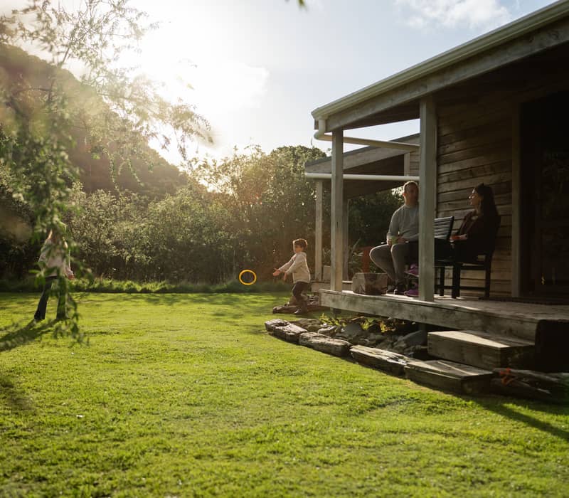 Family relaxing on porch of wooden cabin while kids play on grassy lawn in sunny outdoor setting.