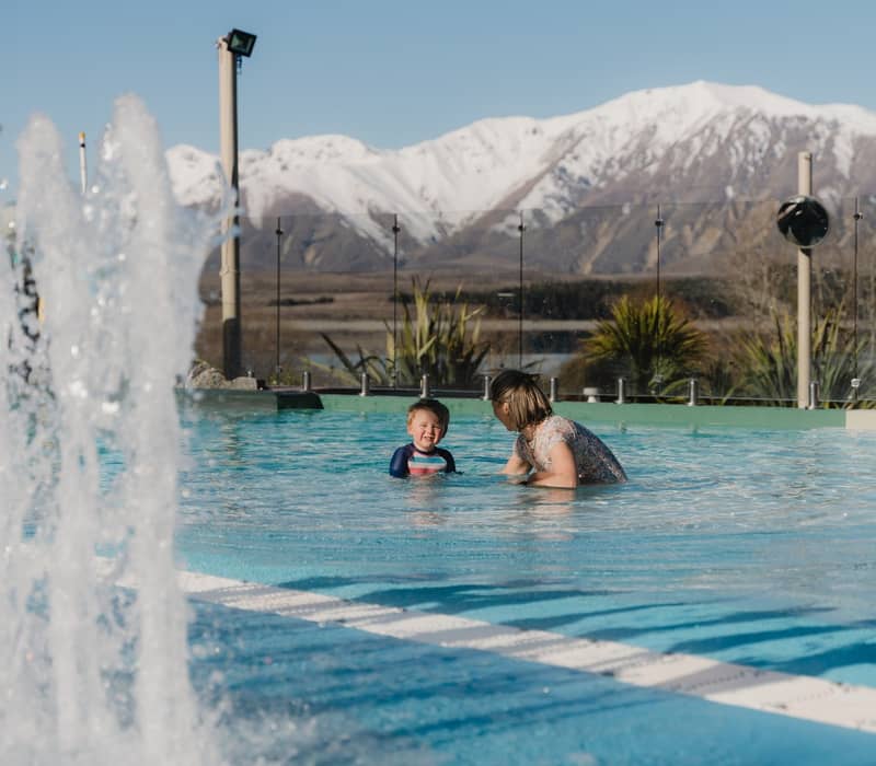 Adult and child swimming in outdoor pool with water fountain and snowy mountains behind. Credit: Tekapo Springs