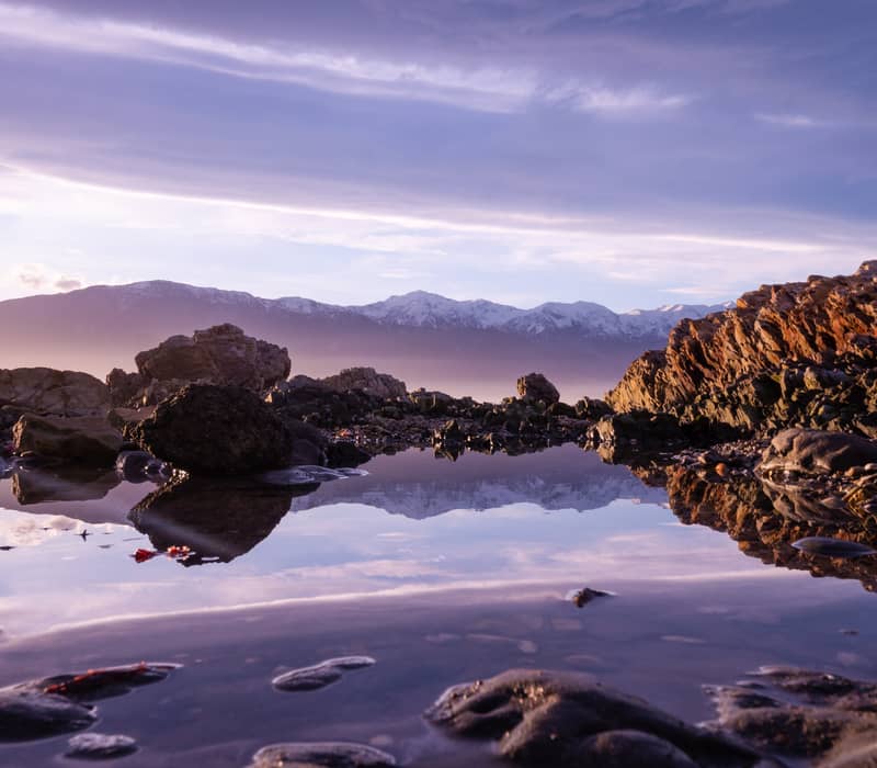 A calm coastal rock pool reflects snow-capped mountains under a soft purple and pink sunset sky.