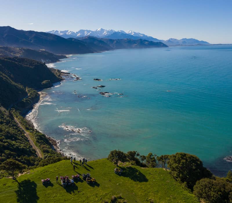 An aerial view of a group of people and quad bikes on a vibrant green grassy cliff overlooking a rugged coastline with turquoise water and distant snow-capped mountains.