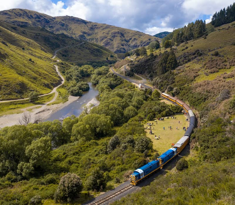 An aerial, high-angle view of a long heritage passenger train with blue and yellow locomotives curving through a deep green valley alongside a winding river, flanked by rugged hills under a blue sky. Credit: Dunedin Railways Ltd