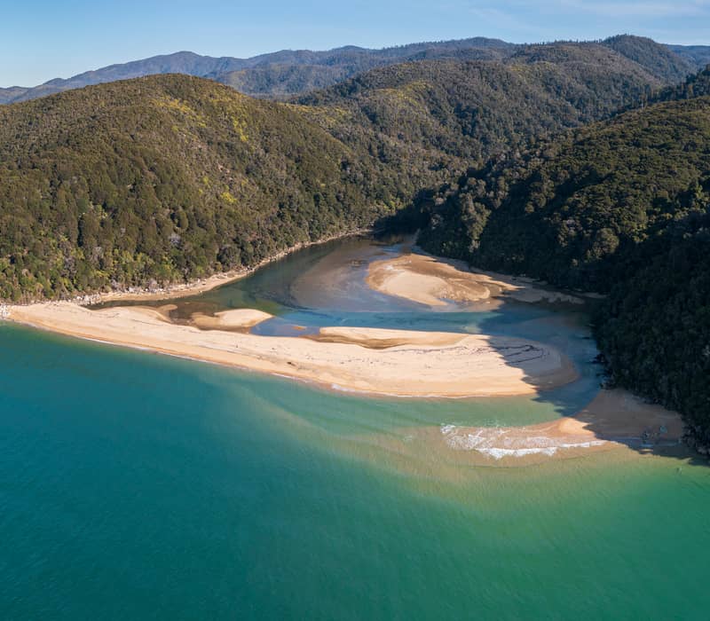 A high-altitude aerial view of a river winding through golden sand to meet a turquoise sea, bordered by lush green forest and a rugged coastline under a clear sky.