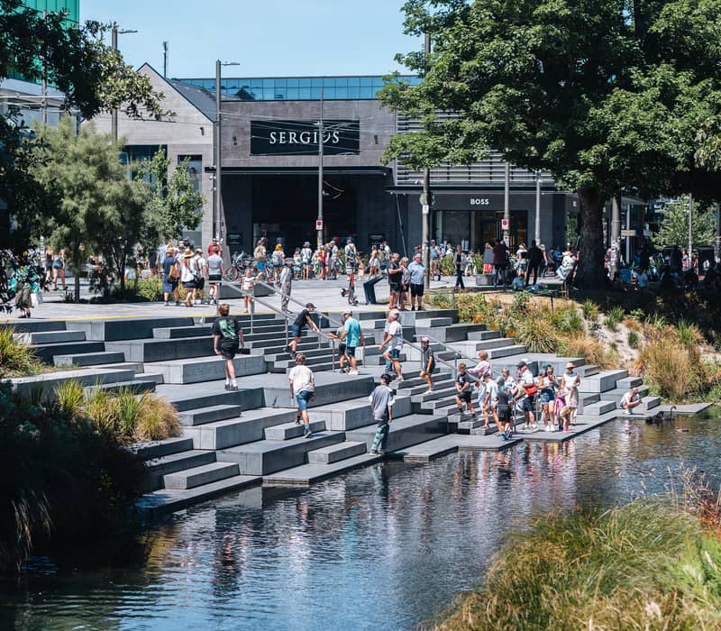 People gathered on steps by the water under trees on a sunny summer day in an urban area.