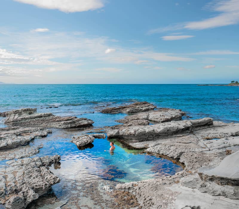 Person bathing in natural rock pool along rocky coastline under blue sky.