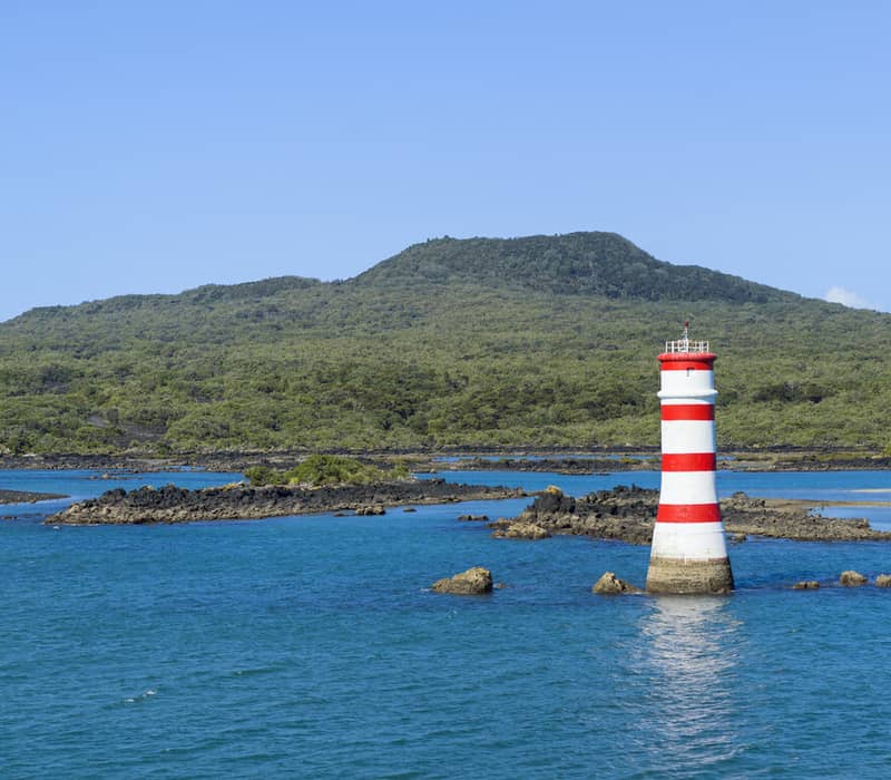 Red and white striped lighthouse in blue water near a green volcanic island with rocky shoreline. Credit: Fullers 360