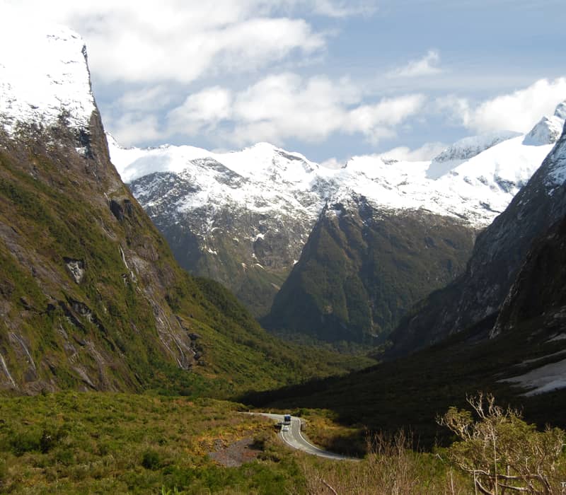 A winding highway snakes through the deep, green Cleddau Valley, surrounded by towering, snow-capped granite peaks and steep mountain walls in Fiordland.