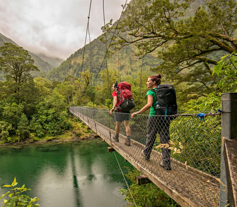 Two hikers with backpacks crossing a suspension bridge over a river in a forested mountain area. Credit: Ultimate Hikes