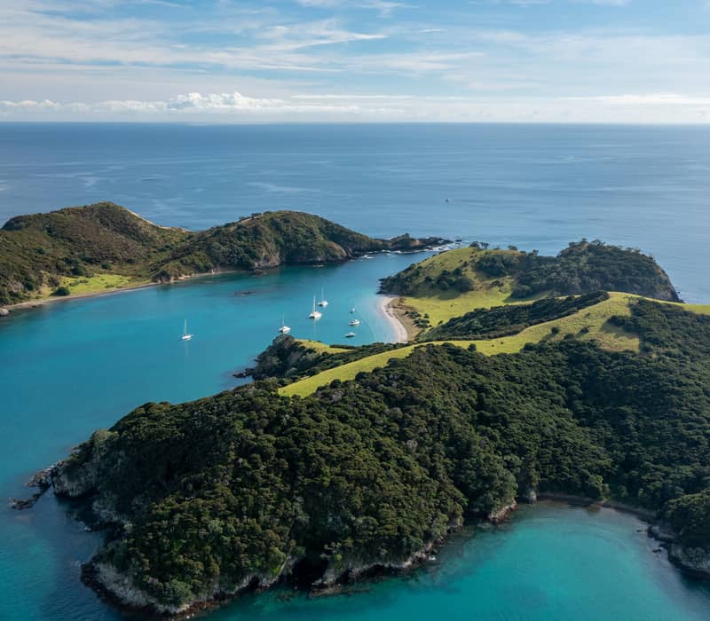 A high-altitude aerial view of Roberton Island in the Bay of Islands, featuring two bright turquoise lagoons separated by a narrow green isthmus with anchored sailboats.