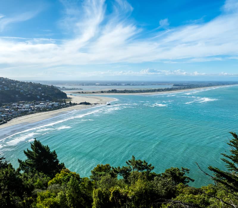 A high-altitude coastal view overlooking a turquoise bay and a long, straight sandy beach stretching into the distance, with a hillside suburb in the foreground under a sky with wispy white clouds.