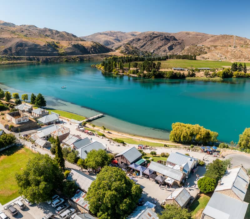 A high-angle aerial view of a historic stone village with market stalls next to a vibrant turquoise lake, with vineyards and rugged dry mountains in the background.