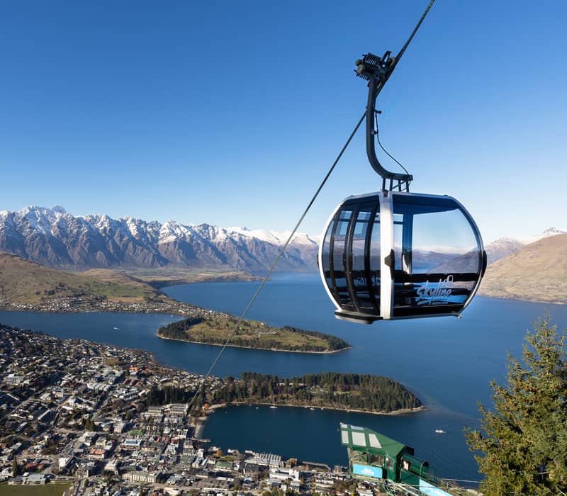 A modern glass gondola cabin suspended high above Queenstown, offering a stunning panoramic view of the blue Lake Wakatipu and the snow-capped Remarkables mountain range. Credit: Skyline
