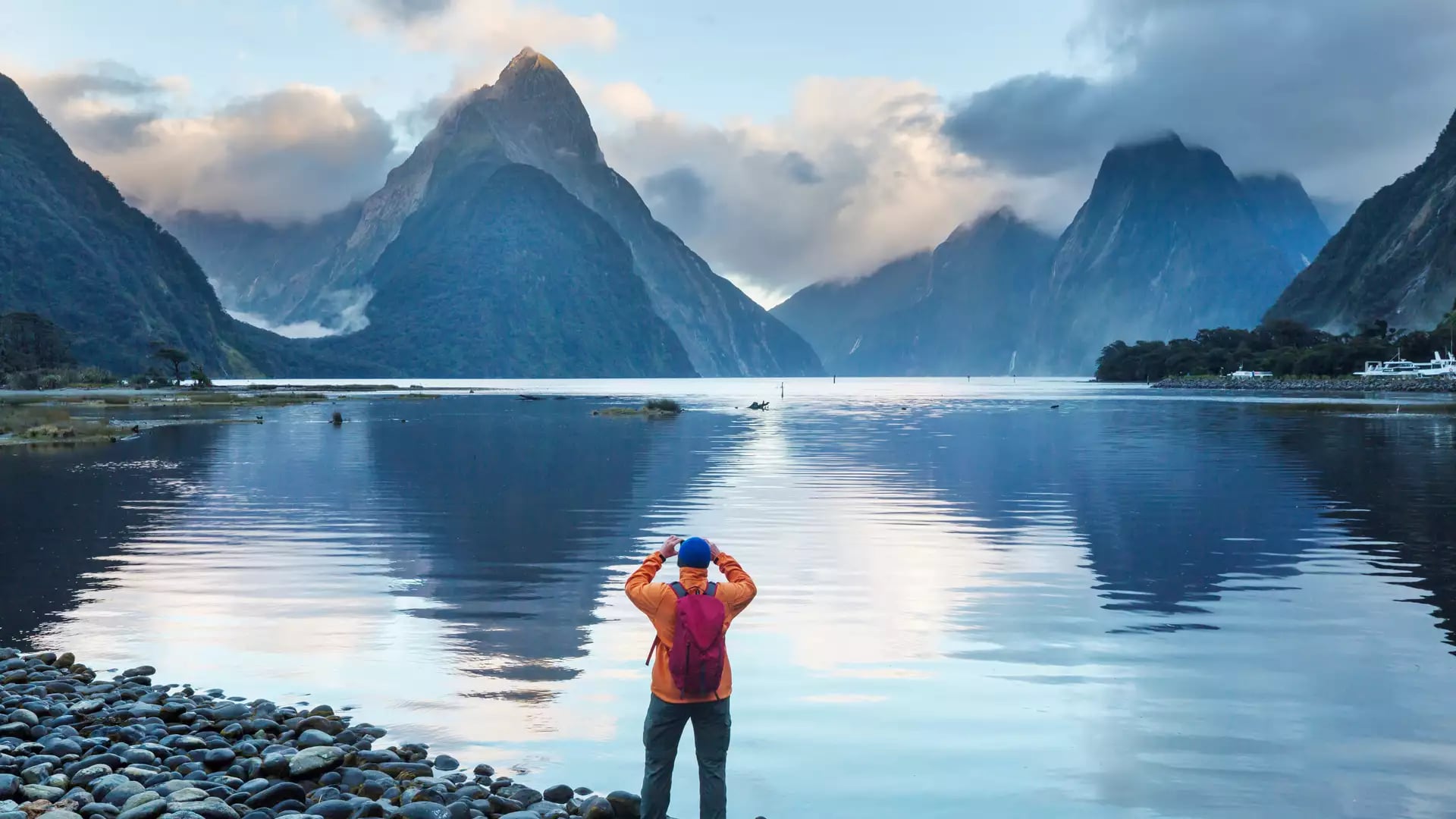 A person in an orange coat looking out over Milford Sound with water and hills in the background Credit: New Zealand Fine Touring - Photographer Unknown