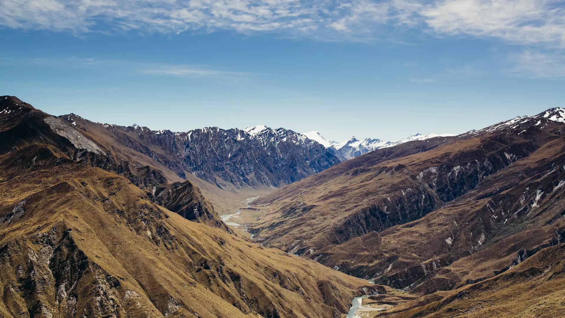 An aerial view over vast, golden-brown mountains with a winding river in a deep valley, leading toward distant snow-capped peaks under a blue sky. Credit: Wanaka Tourism