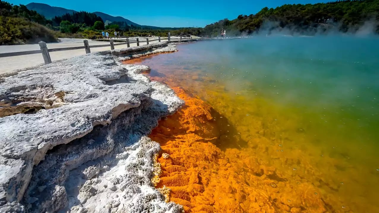 A close-up view of the vibrant orange mineral edge and turquoise steaming water of the Champagne Pool, with a wooden boardwalk and visitors in the background. Credit: Rotorua NZ - Media Library