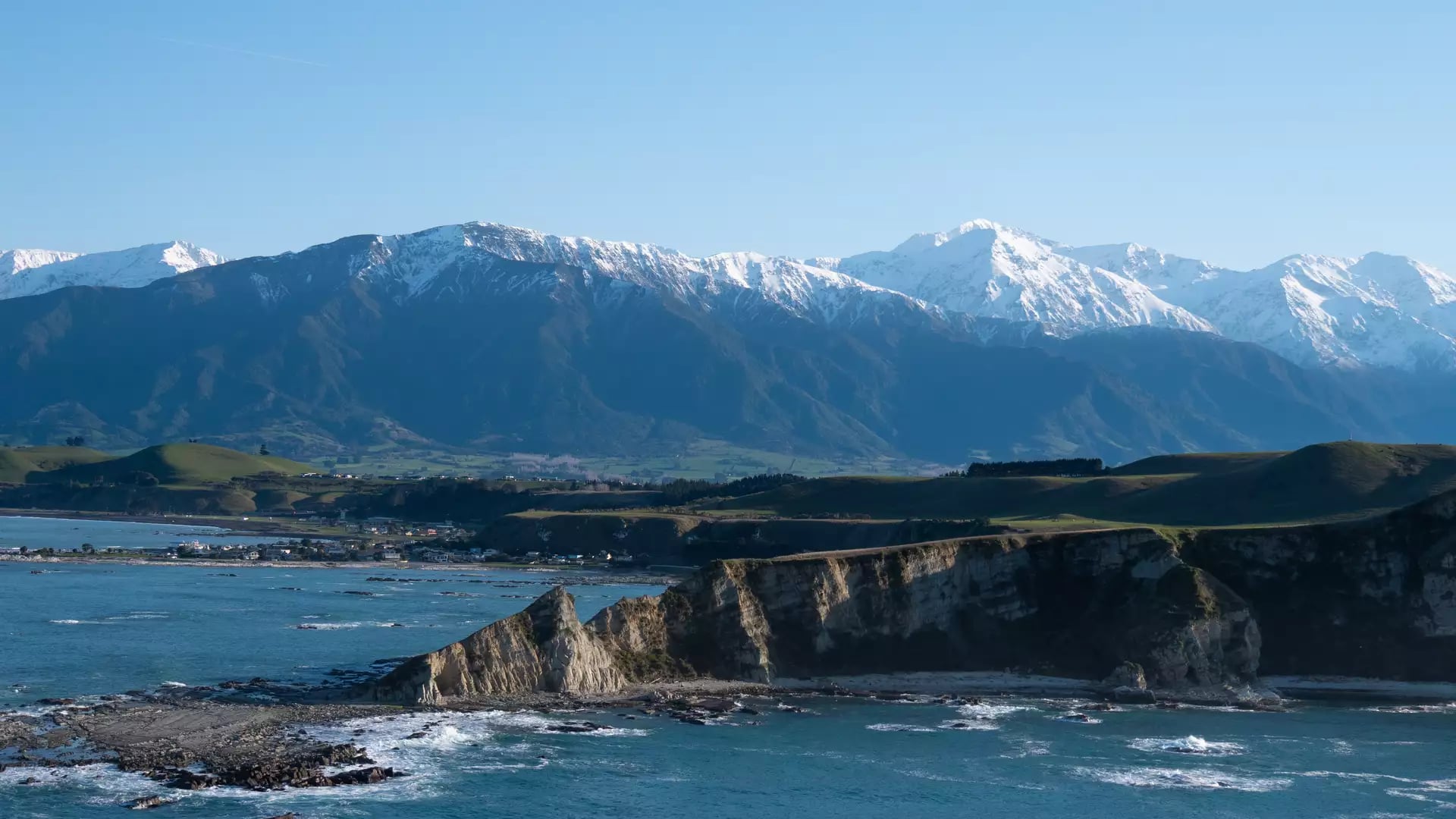 A high-angle coastal view featuring sharp white limestone cliffs jutting into a turquoise ocean, with rolling green hills and a vast range of snow-capped mountains in the background under a clear blue sky.