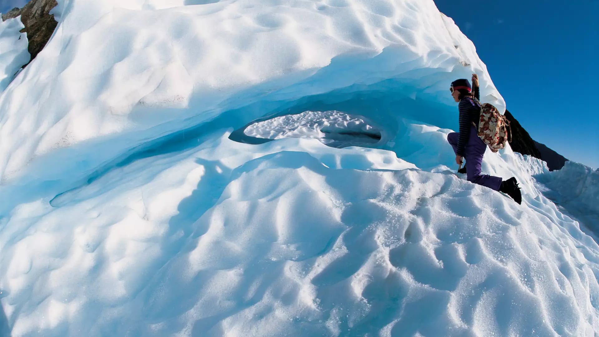 A hiker in purple gear and a backpack explores a glowing blue ice cave and dramatic ridges on the surface of Fox Glacier. Credit: Fine Tours Group - Client image