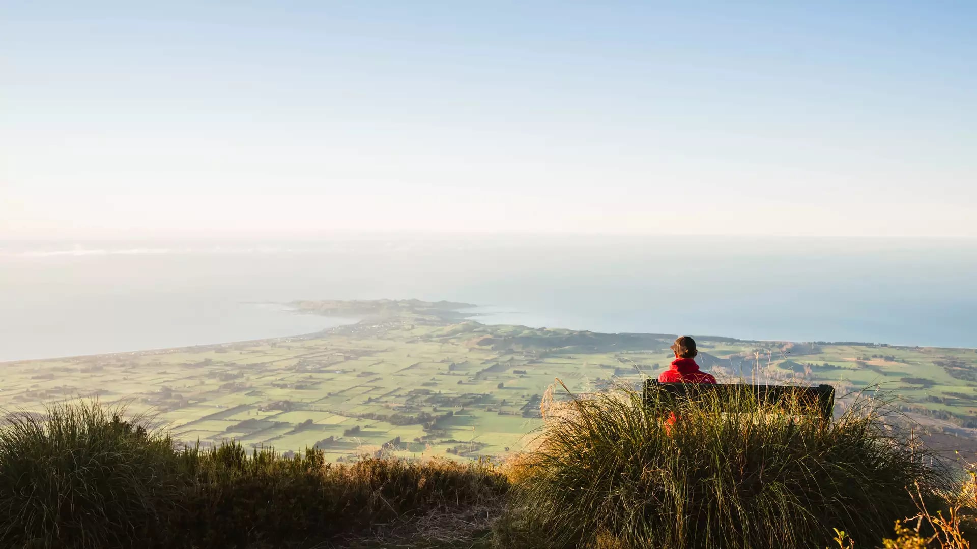 Person in red jacket sitting on bench on Mt Fyffe overlooking coastal landscape at sunset
