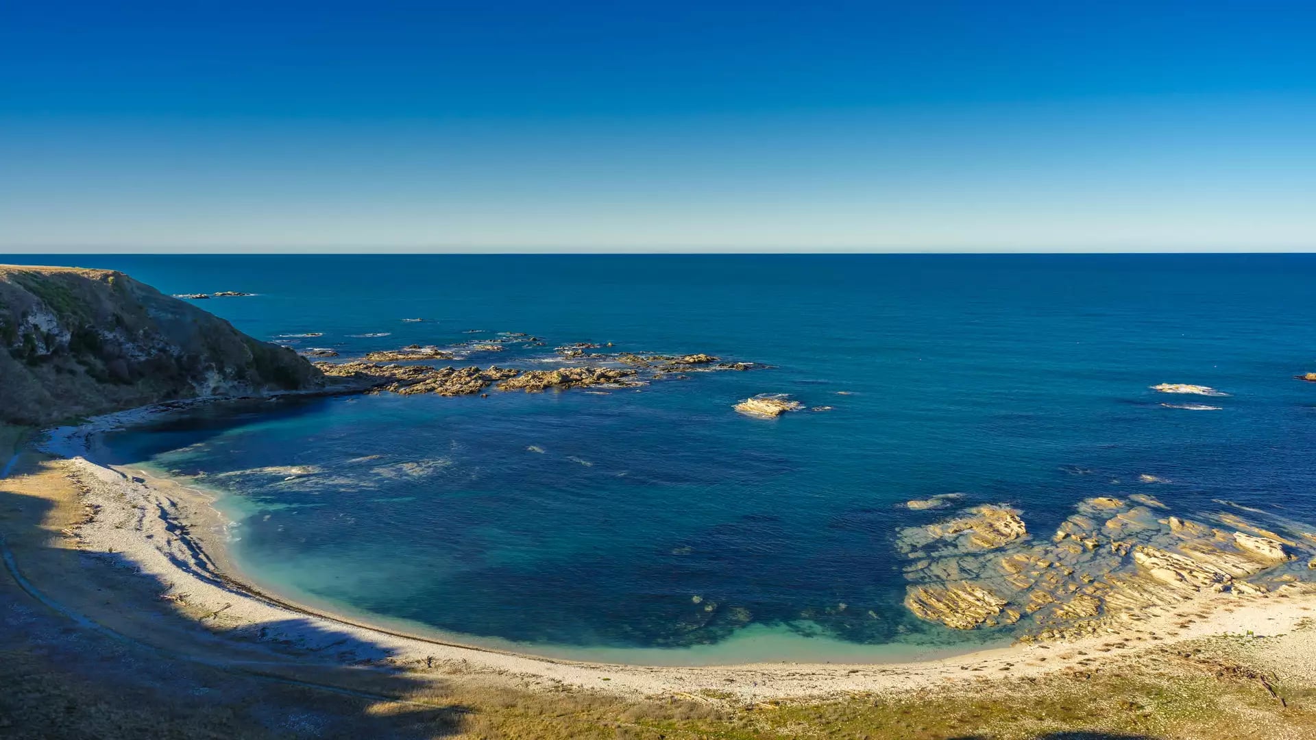 Clear blue ocean and rocky beach forming a curved bay under sunny blue sky. Credit: New Zealand Fine Touring