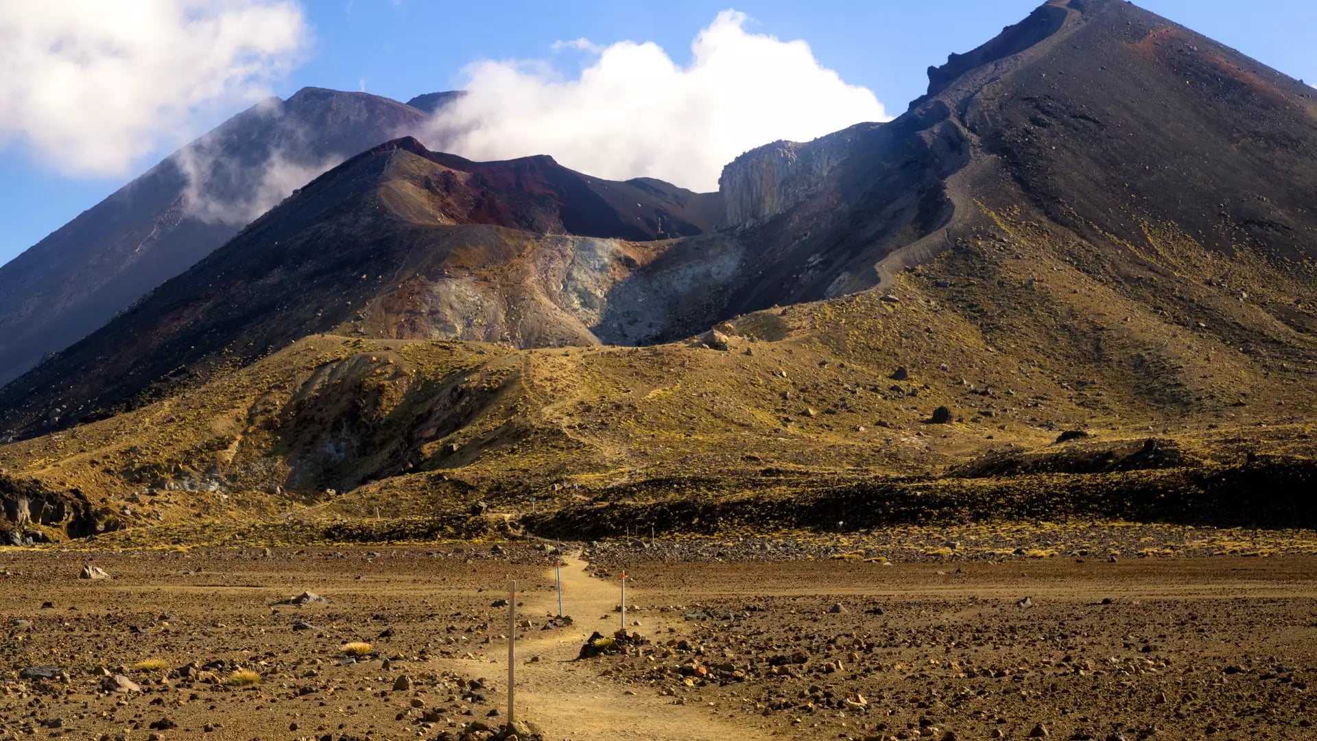 Volcanic mountain Mt Ngauruhoe with hiking trail in foreground under blue sky with clouds.