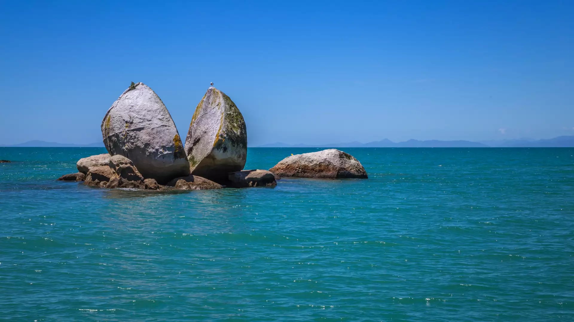 A large split granite boulder in the ocean under a clear blue sky. Credit: New Zealand Fine Touring / Photographer Unknown