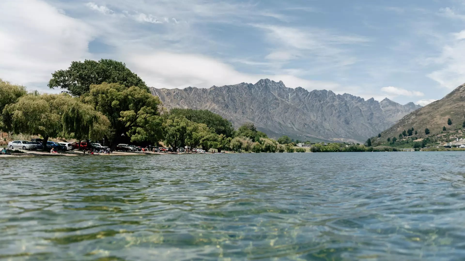 A wide, low-angle shot from the clear, rippling waters of Frankton Beach, with leafy willow trees lining the shore, parked cars, and the jagged, snow-capped Remarkables mountain range in the background.