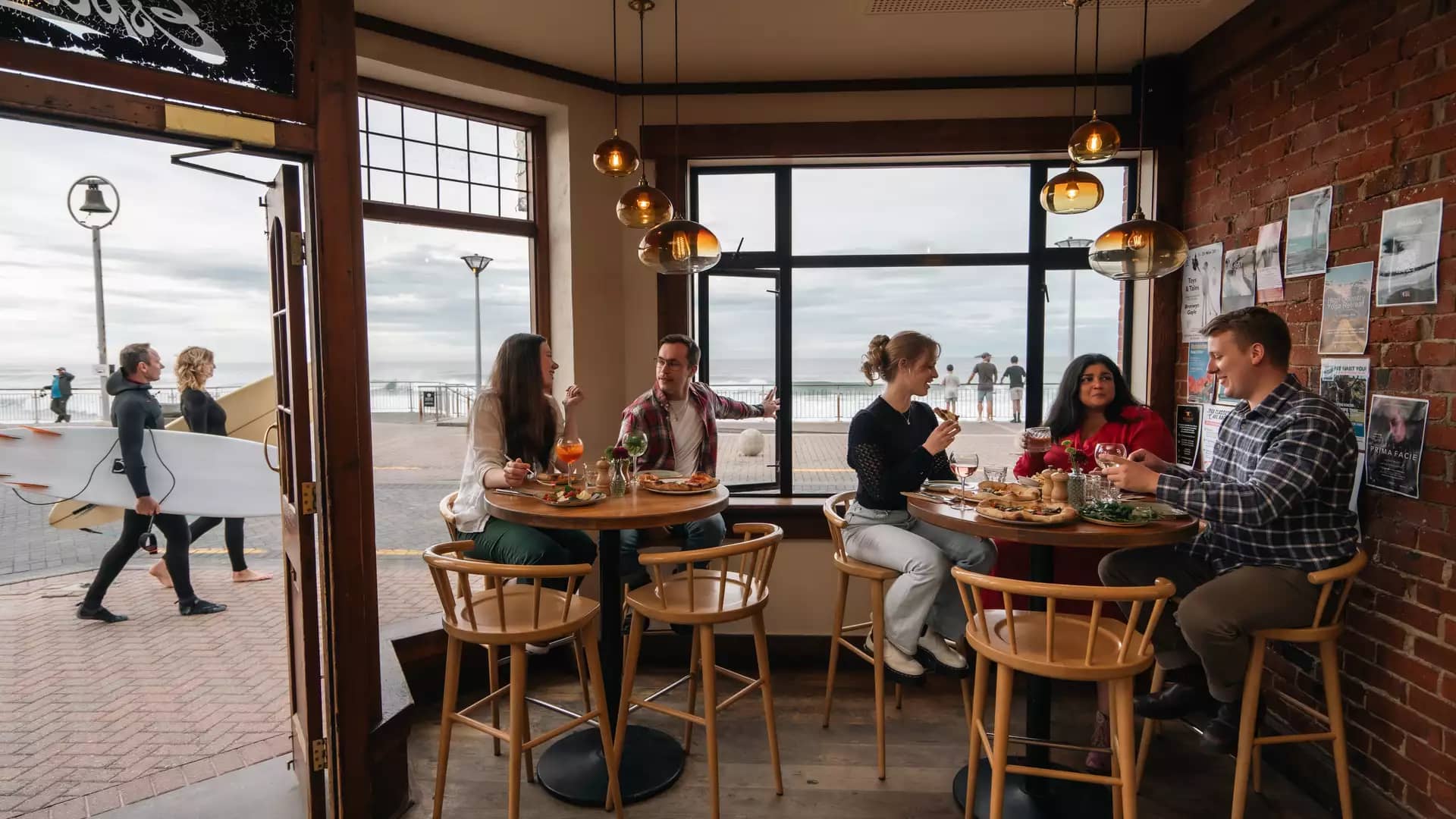 People dining inside a seaside café with ocean view and surfers walking outside the door.