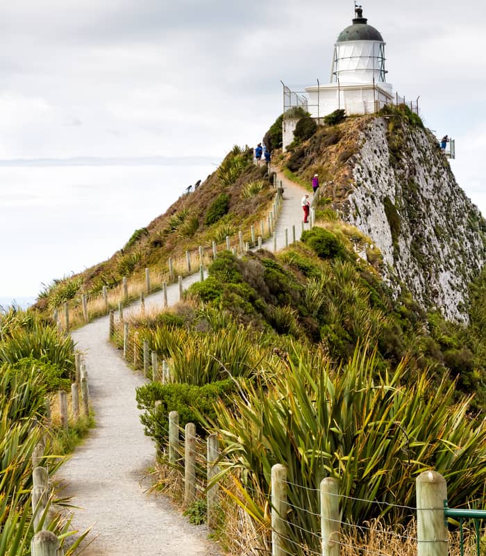 Path leading up to Nugget Point Lighthouse on a cliff with tourists walking and cloudy sky.